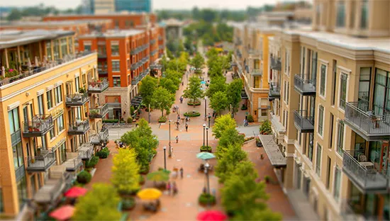 A tilt-shift view down a tree-lined main street with residential balconies, sidewalk cafés, umbrellas, and people walking the central pedestrian spine.